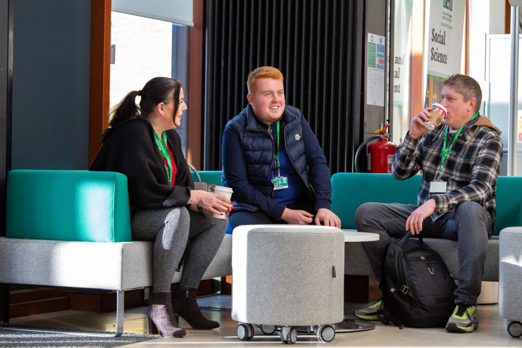 3 students sit together chatting. The middle student is in focus, with the students to the left and right looking towards them and listening intently.