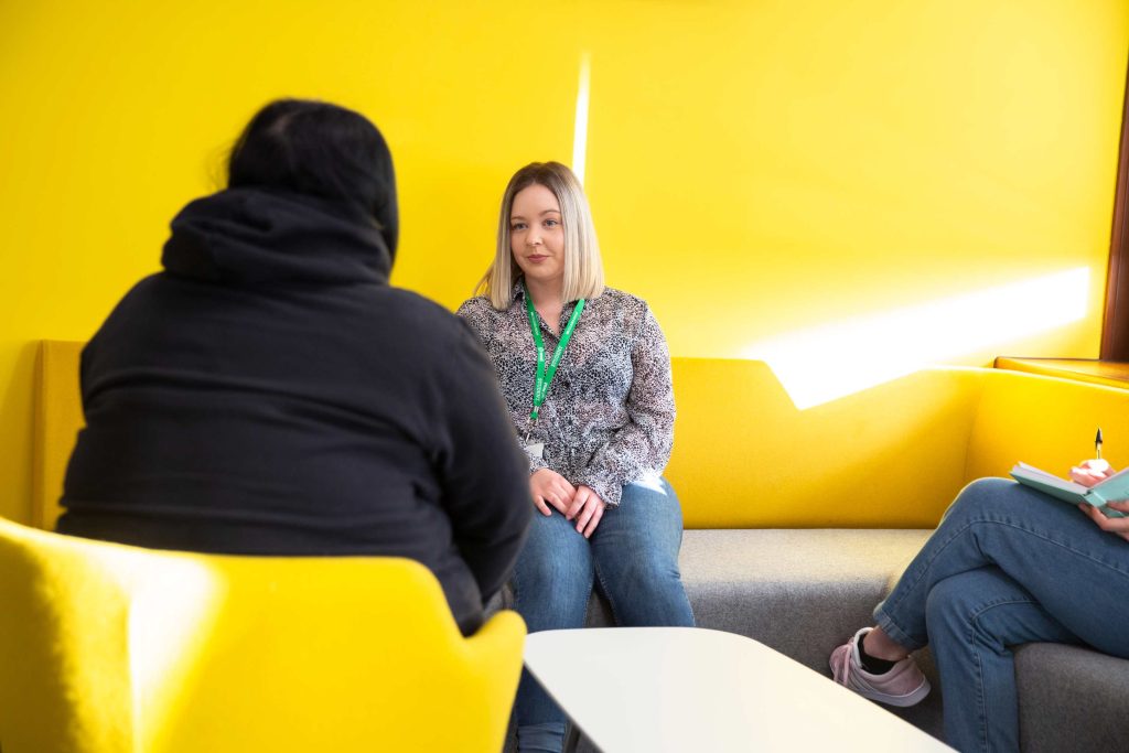 A student sits smiling chatting to another person. They are sat in a brightly lit environment with a vibrant yellow wall dominating the background behind them