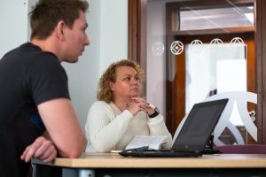 2 students sit at a table looking intently as if deep in thought. A laptop is on the table in front of the student closest to us.