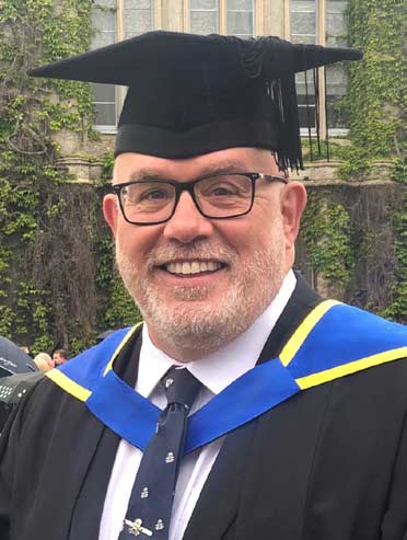 Andy posing wearing graduate mortar board cap, and gown with a blue and yellow trimmed hood around neck and shoulders