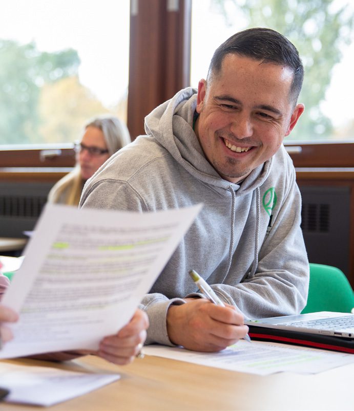 A student wearing a grey UCNL branded hoodie smiles at a table, pen in hand while chatting with fellow students who are outside the image borders