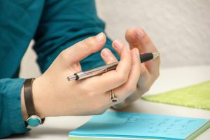 A close up image of a person's hands, with a pen resting across them. Their hands are hovering over a small note pad full of some notes.